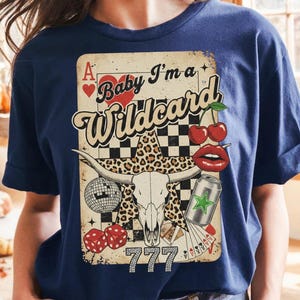 May include: Navy blue t-shirt featuring a graphic with a playing card design. The design includes a bull skull, cherries, dice, and the text "Baby I'm a Wildcard" with the number "777".