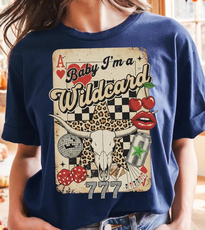 May include: Navy blue t-shirt featuring a graphic with a playing card design. The design includes a bull skull, cherries, dice, and the text "Baby I'm a Wildcard" with the number "777".