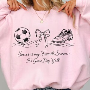 May include: Light pink sweatshirt with a soccer ball, bow, and soccer cleat graphic. The text "Soccer is my Favorite Season It's Game Day Y'all" is printed below the graphics. The sweatshirt is a casual, sporty style.