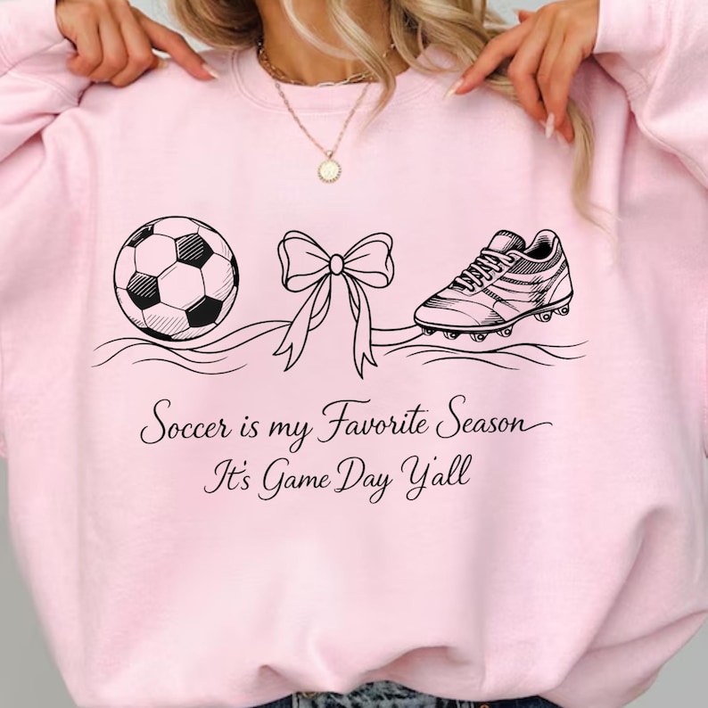 May include: Light pink sweatshirt with a soccer ball, bow, and soccer cleat graphic. The text "Soccer is my Favorite Season It's Game Day Y'all" is printed below the graphics. The sweatshirt is a casual, sporty style.