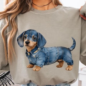 May include: A light gray long-sleeve shirt featuring a dachshund wearing a denim jacket and overalls. The dog has brown and tan fur with blue denim clothing. The shirt is a neutral color.
