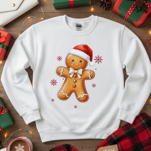 May include: White sweatshirt featuring a gingerbread man wearing a Santa hat and bow tie. The gingerbread man has a smiling face and is surrounded by snowflakes. The sweatshirt is a festive holiday design.