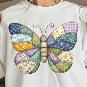 May include: White sweatshirt featuring a large, colorful butterfly design. The butterfly is made of patchwork fabric in various patterns and colors, including blue, purple, yellow, and green. The design has a stitched, quilted appearance.
