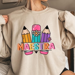 May include: Beige sweatshirt with a colorful graphic design featuring three pencils, an eraser, and the word "MAESTRA" in a playful font. The design includes orange, yellow, teal, purple, pink, and white colors. A fun and educational theme.