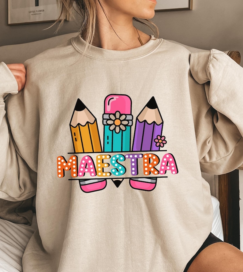 May include: Beige sweatshirt with a colorful graphic design featuring three pencils, an eraser, and the word "MAESTRA" in a playful font. The design includes orange, yellow, teal, purple, pink, and white colors. A fun and educational theme.