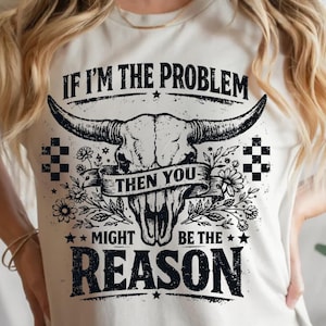 May include: Cream-colored t-shirt featuring a black and white graphic of a bull skull with large horns, floral accents, and the text "IF I'M THE PROBLEM THEN YOU MIGHT BE THE REASON."
