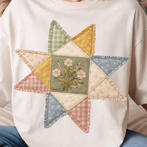 May include: Cream-colored t-shirt featuring a large, colorful star design. The star is composed of various fabric patches in pastel shades of pink, yellow, blue, and green, with a central square embroidered with flowers.