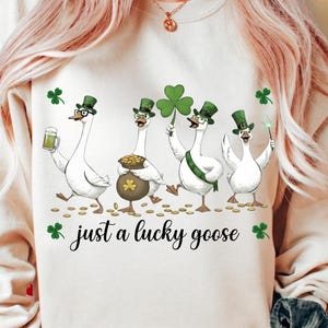 Puede incluir: Sudadera color crema con un gr&aacute;fico del D&iacute;a de San Patricio. El dise&ntilde;o presenta cuatro gansos de dibujos animados con sombreros y accesorios verdes, sosteniendo una cerveza, una olla de oro, un tr&eacute;bol y una varita. El texto "just a lucky goose" est&aacute; debajo.