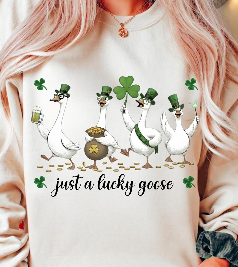 Puede incluir: Sudadera color crema con un gr&aacute;fico del D&iacute;a de San Patricio. El dise&ntilde;o presenta cuatro gansos de dibujos animados con sombreros y accesorios verdes, sosteniendo una cerveza, una olla de oro, un tr&eacute;bol y una varita. El texto "just a lucky goose" est&aacute; debajo.