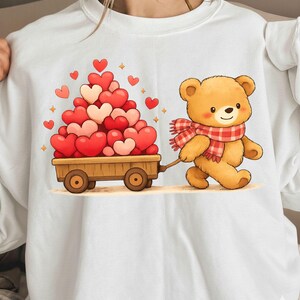 May include: White sweatshirt with a cartoon teddy bear pulling a wooden cart laden with red and pink hearts. The bear is wearing a red and white checked scarf. A cute design for Valentine's Day or a special gift.