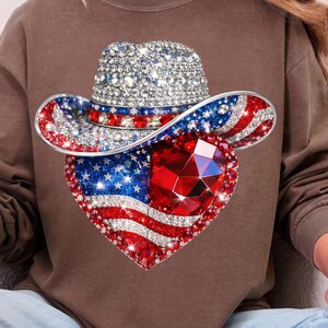 May include: A bedazzled cowboy hat with a heart-shaped design. The hat features a silver crown and a brim adorned with red, white, and blue crystals, forming the American flag. A large, faceted red gem is at the heart's center.
