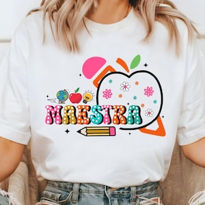 May include: White t-shirt with a colorful graphic design. The design includes the word "MAESTRA" in a retro font, an apple, flowers, a pencil, a globe, and a lightbulb. The design is in shades of pink, orange, blue, green, and yellow.