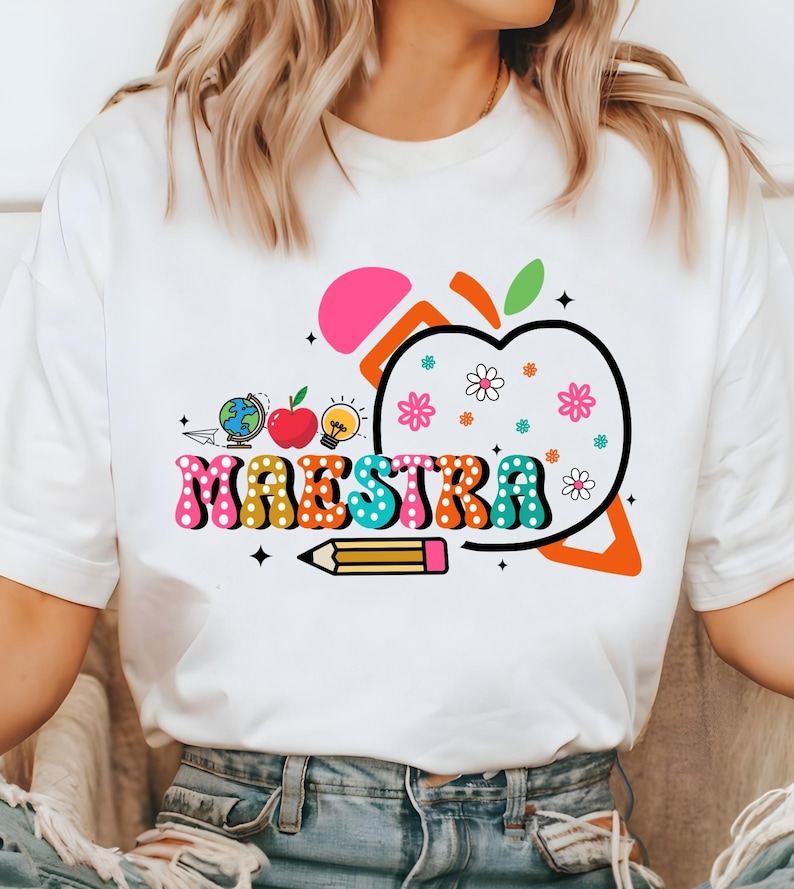 May include: White t-shirt with a colorful graphic design. The design includes the word "MAESTRA" in a retro font, an apple, flowers, a pencil, a globe, and a lightbulb. The design is in shades of pink, orange, blue, green, and yellow.