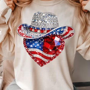 May include: A cream-colored long-sleeve shirt featuring a rhinestone-studded cowboy hat and a heart-shaped design with a red, white, and blue American flag motif. The hat and heart are embellished with sparkling rhinestones.