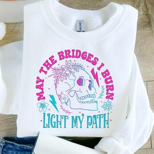 May include: White sweatshirt with a graphic design featuring a skull, flowers, and lightning bolts. The text reads "MAY THE BRIDGES I BURN LIGHT MY PATH" in pink and teal lettering. The sweatshirt is folded.
