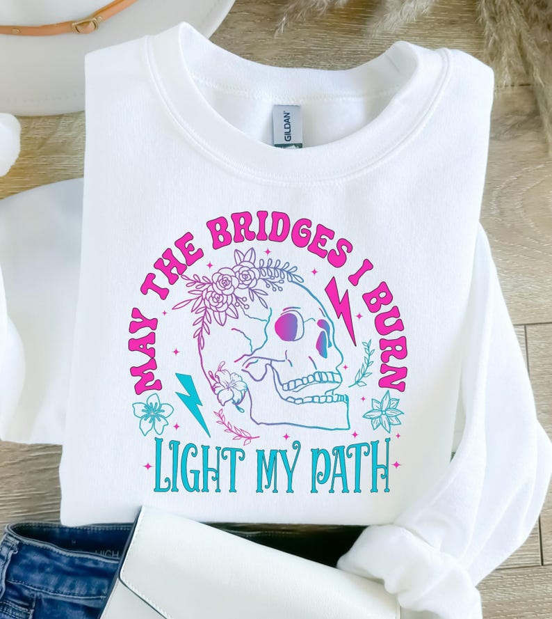 May include: White sweatshirt with a graphic design featuring a skull, flowers, and lightning bolts. The text reads "MAY THE BRIDGES I BURN LIGHT MY PATH" in pink and teal lettering. The sweatshirt is folded.