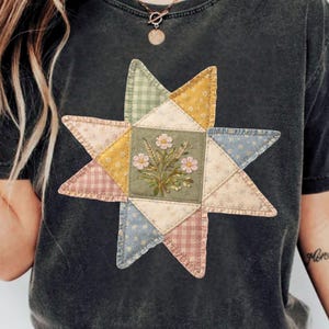 May include: Dark gray t-shirt featuring a colorful star-shaped quilt design. The quilt has a central square with floral embroidery and is surrounded by triangles in various pastel patterns. The shirt is made of a soft material.