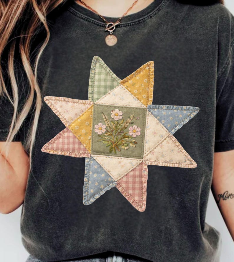 May include: Dark gray t-shirt featuring a colorful star-shaped quilt design. The quilt has a central square with floral embroidery and is surrounded by triangles in various pastel patterns. The shirt is made of a soft material.