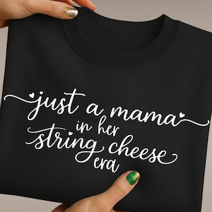 May include: Black crew neck sweatshirt with white cursive text that reads "just a mama in her string cheese era". The text is centered on the front of the shirt. The sweatshirt is held by two hands.