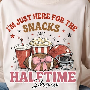 May include: A cream-coloured t-shirt with a graphic design featuring a football, popcorn, a drink, and a helmet. The text reads "I'M JUST HERE FOR THE SNACKS AND HALFTIME SHOW."