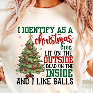 Puede incluir: Camiseta color crema con un gráfico de árbol de Navidad y el texto "I IDENTIFY AS A christmas tree LIT ON THE OUTSIDE DEAD ON THE INSIDE AND I LIKE BALLS". El gráfico presenta un árbol de Navidad decorado con adornos rojos.