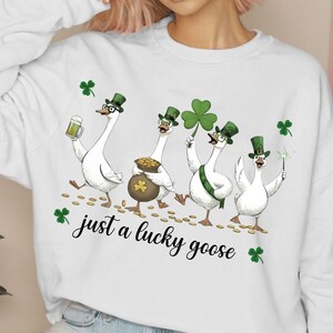 Puede incluir: Sudadera blanca con un dise&ntilde;o del D&iacute;a de San Patricio. Cuatro gansos de dibujos animados llevan sombreros verdes y sostienen una cerveza, una olla de oro, un tr&eacute;bol y una bengala. El texto "just a lucky goose" est&aacute; escrito en cursiva.