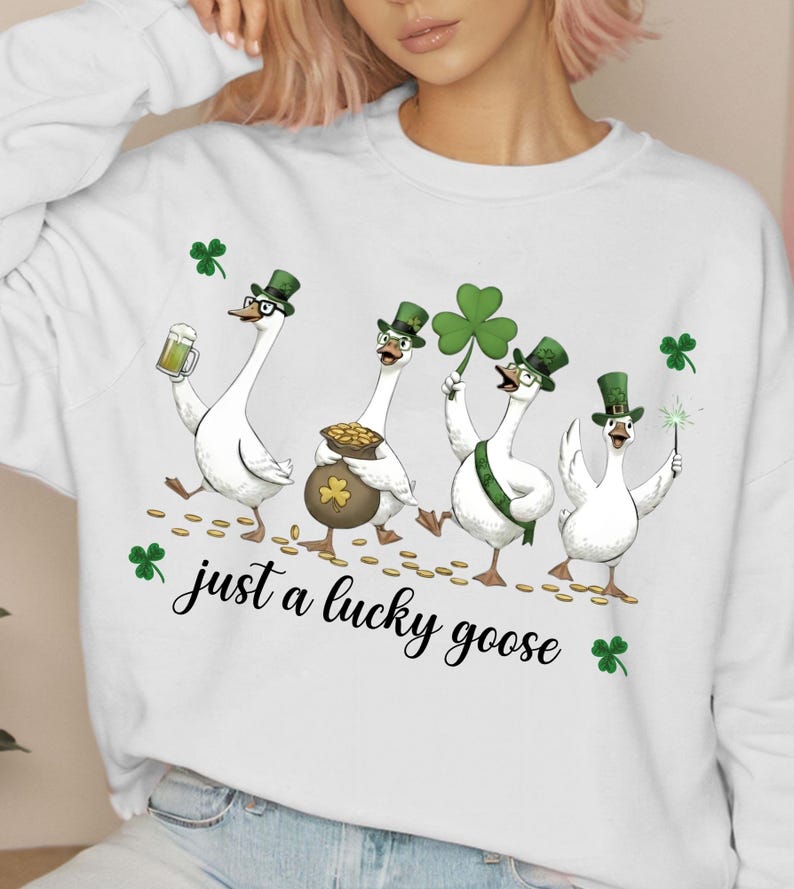 Puede incluir: Sudadera blanca con un dise&ntilde;o del D&iacute;a de San Patricio. Cuatro gansos de dibujos animados llevan sombreros verdes y sostienen una cerveza, una olla de oro, un tr&eacute;bol y una bengala. El texto "just a lucky goose" est&aacute; escrito en cursiva.