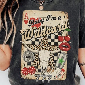 May include: Dark gray t-shirt with a graphic featuring a playing card design. The design includes a heart, cherries, lips, a disco ball, dice, a skull, and the text "Baby I'm a Wildcard" and "777".