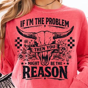 May include: A coral-colored long-sleeve sweatshirt with a graphic design. The design features a bull skull with large horns, floral accents, and the text "IF I'M THE PROBLEM THEN YOU MIGHT BE THE REASON."