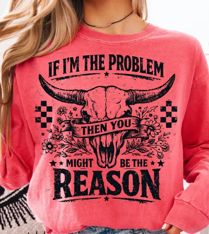 May include: A coral-colored long-sleeve sweatshirt with a graphic design. The design features a bull skull with large horns, floral accents, and the text "IF I'M THE PROBLEM THEN YOU MIGHT BE THE REASON."