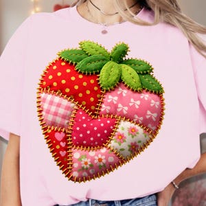 Puede incluir: Una camiseta rosa claro con un gran diseño de fresa en patchwork. La fresa está compuesta por varios patrones de tela, incluyendo lunares rojos, lazos rosas y estampados florales. El diseño está delineado con costuras amarillas y tiene hojas verdes de fieltro.