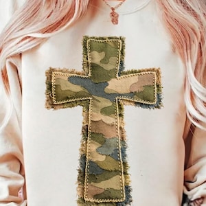 Puede incluir: Sudadera de color crema con una gran cruz aplicada. La cruz está hecha de tela de camuflaje en tonos verdes, marrones y azules, con un borde deshilachado. El diseño está centrado en la parte delantera de la prenda.