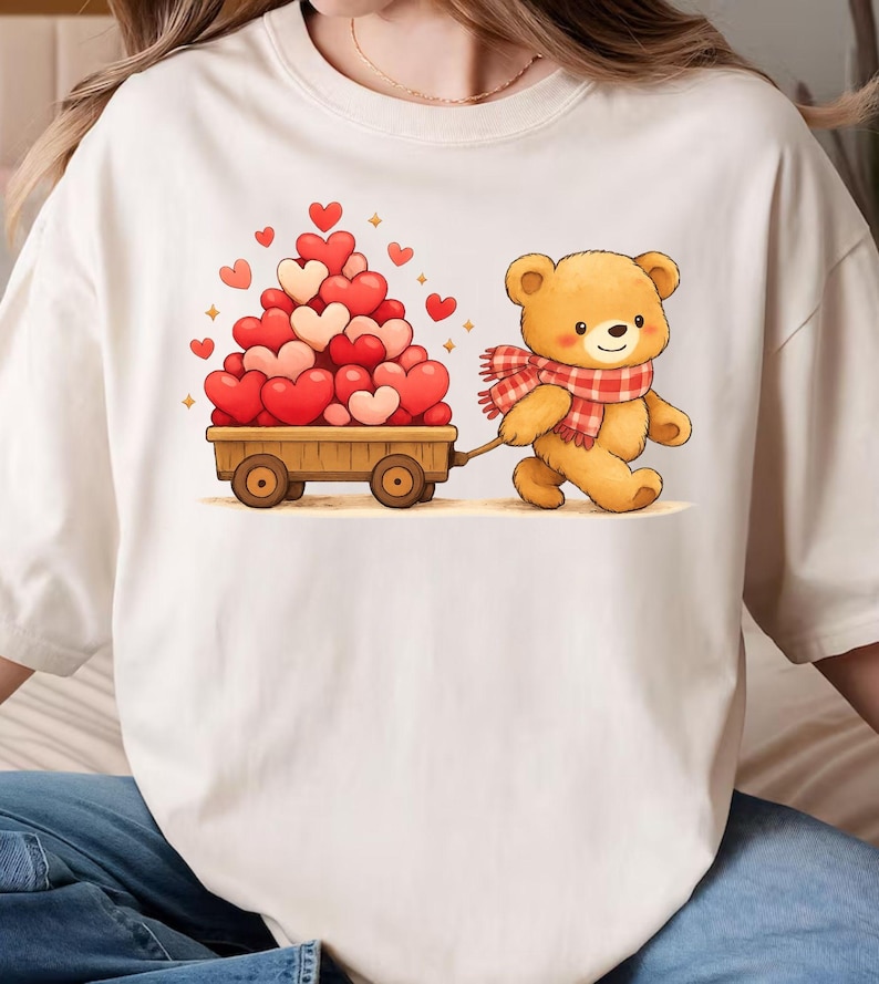 May include: A cream t-shirt with a cartoon teddy bear wearing a red and white scarf, pulling a wooden cart filled with red and pink hearts. The design includes small heart and star accents.