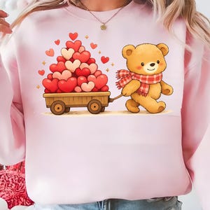 May include: Pale pink sweatshirt with a cartoon teddy bear pulling a wooden cart piled high with red and pink hearts. The bear wears a red and white checked scarf. A charming design for Valentine's Day or a romantic gift.