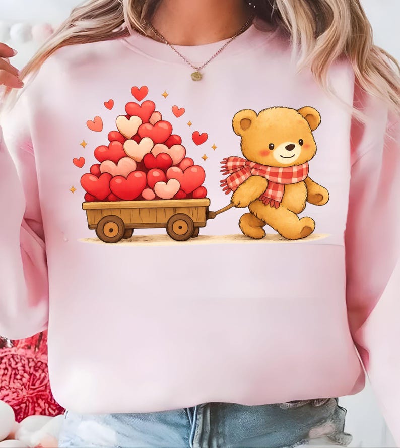 May include: Pale pink sweatshirt with a cartoon teddy bear pulling a wooden cart piled high with red and pink hearts. The bear wears a red and white checked scarf. A charming design for Valentine's Day or a romantic gift.