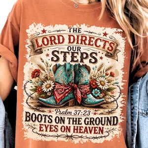 May include: Burnt orange t-shirt featuring a graphic design with turquoise cowboy boots, a red bandana, and floral accents. The text reads "The Lord Directs Our Steps" and "Boots on the Ground Eyes on Heaven" with a Bible verse reference.