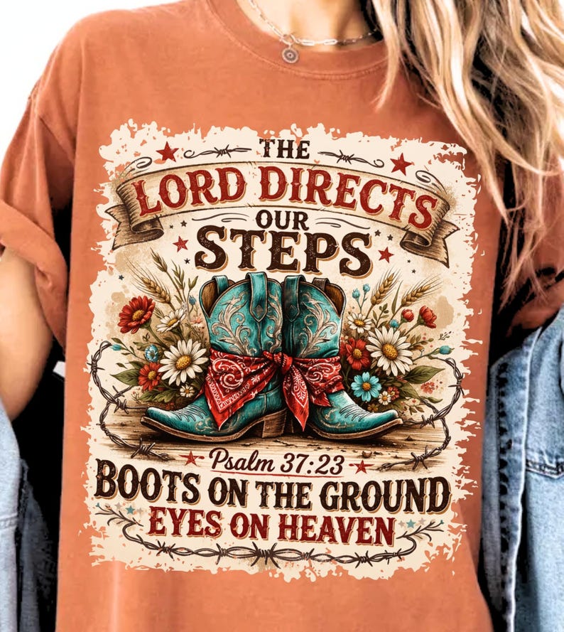 May include: Burnt orange t-shirt featuring a graphic design with turquoise cowboy boots, a red bandana, and floral accents. The text reads "The Lord Directs Our Steps" and "Boots on the Ground Eyes on Heaven" with a Bible verse reference.