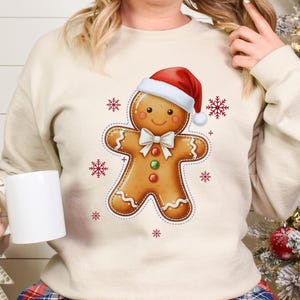May include: A cream-colored sweatshirt featuring a gingerbread man graphic wearing a Santa hat and bow tie. Red snowflakes are scattered around the gingerbread man. The person is holding a white mug.