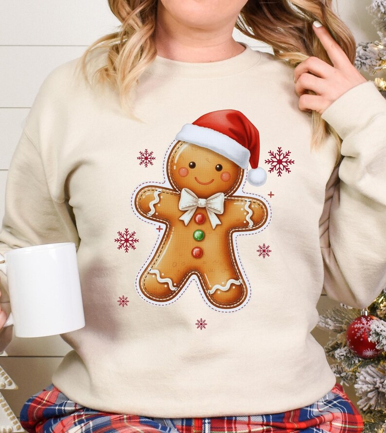 May include: A cream-colored sweatshirt featuring a gingerbread man graphic wearing a Santa hat and bow tie. Red snowflakes are scattered around the gingerbread man. The person is holding a white mug.