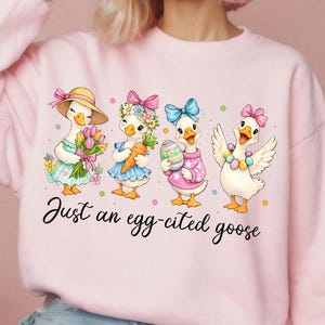 May include: Pink sweatshirt featuring a whimsical illustration of four geese dressed for Easter. Each goose is adorned with accessories like hats, bows, and holding Easter-themed items. The text "Just an egg-cited goose" is written in cursive.