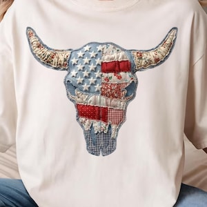 May include: White t-shirt featuring a bull skull design. The skull is crafted from denim, red, white, and floral patterned fabric, with white stars and a patchwork design. The horns are adorned with floral fabric.