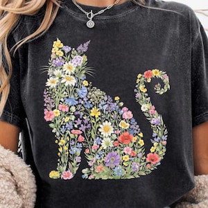 May include: Dark gray t-shirt featuring a floral cat design. The cat silhouette is filled with colorful flowers in shades of yellow, white, purple, blue, and red. The shirt has short sleeves and a crew neck.