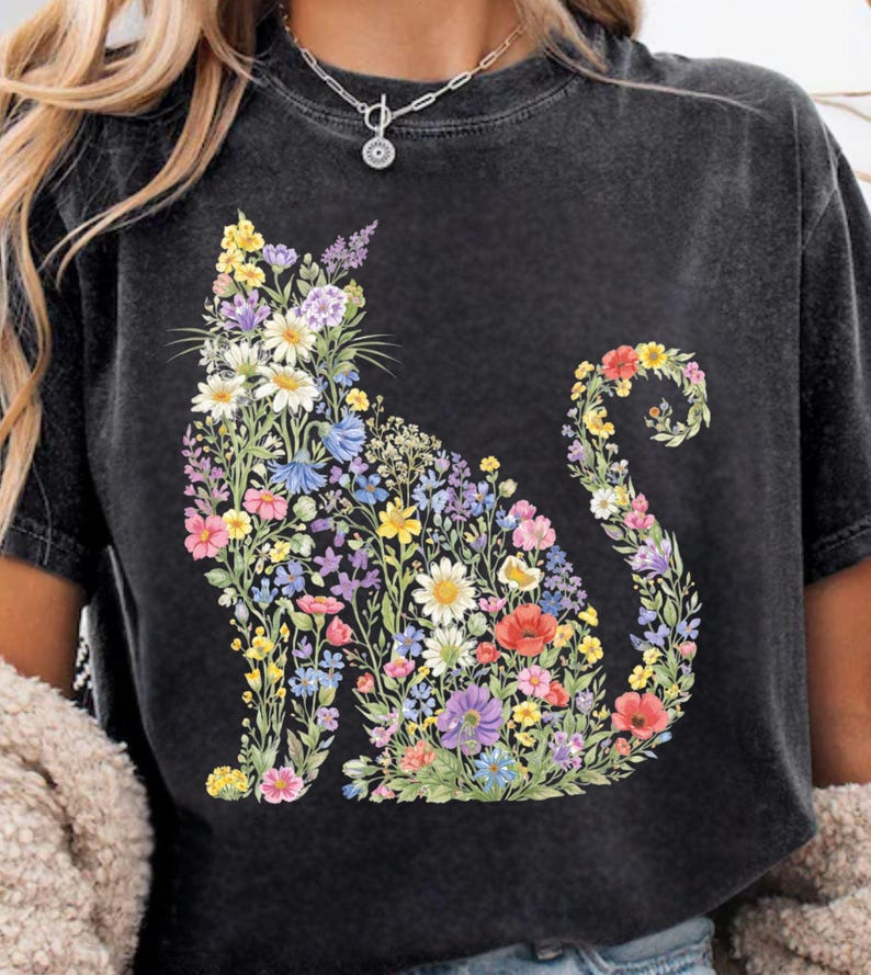 May include: Dark gray t-shirt featuring a floral cat design. The cat silhouette is filled with colorful flowers in shades of yellow, white, purple, blue, and red. The shirt has short sleeves and a crew neck.
