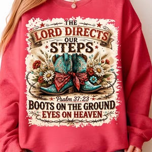 May include: Red sweatshirt with a graphic featuring turquoise cowboy boots, floral accents, and the text "The Lord Directs Our Steps." The design includes the Bible verse Psalm 37:23 and the phrase "Boots on the Ground, Eyes on Heaven."