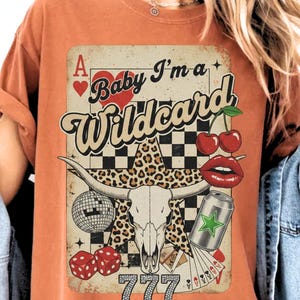 May include: Orange t-shirt featuring a graphic with a vintage design. The design includes a heart, cherries, dice, a disco ball, a can, a skull, and the text "Baby I'm a Wildcard" and "777".
