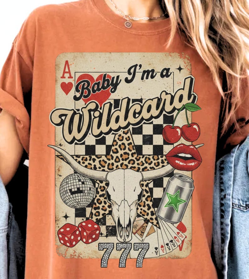 May include: Orange t-shirt featuring a graphic with a vintage design. The design includes a heart, cherries, dice, a disco ball, a can, a skull, and the text "Baby I'm a Wildcard" and "777".