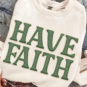 May include: White sweatshirt with the words "HAVE FAITH" in green, textured lettering. The letters have a light beige outline. The sweatshirt has a crew neck and long sleeves. The background includes a denim jacket.