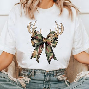 May include: White t-shirt featuring a camouflage bow design with deer antlers. The bow has shades of green, brown, and tan. The antlers are a light brown color. The shirt is a casual style.