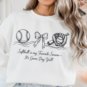 May include: White sweatshirt with a black graphic of a softball, bow, and glove with a softball inside. The text reads "Softball is my Favorite Season, It's Game Day Y'all."