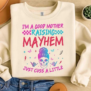 May include: Cream-colored sweatshirt with the text "I'M A GOOD MOTHER RAISING MAYHEM JUST CUSS A LITTLE" in pink and blue. The design includes a skull with a bun, butterflies, and hand gestures.